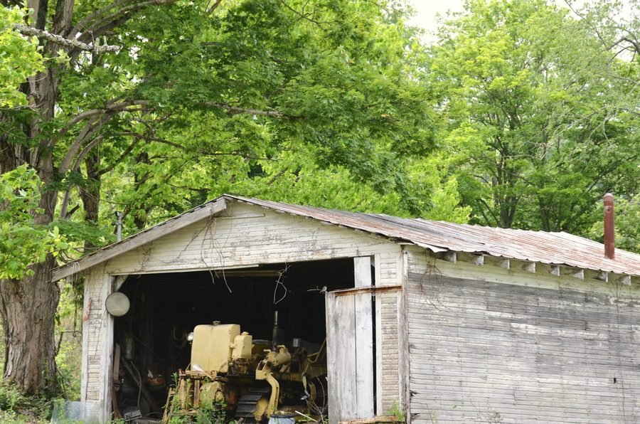 A weathered wooden machine shed with its doors open and yellow equipment inside, under tall green trees.