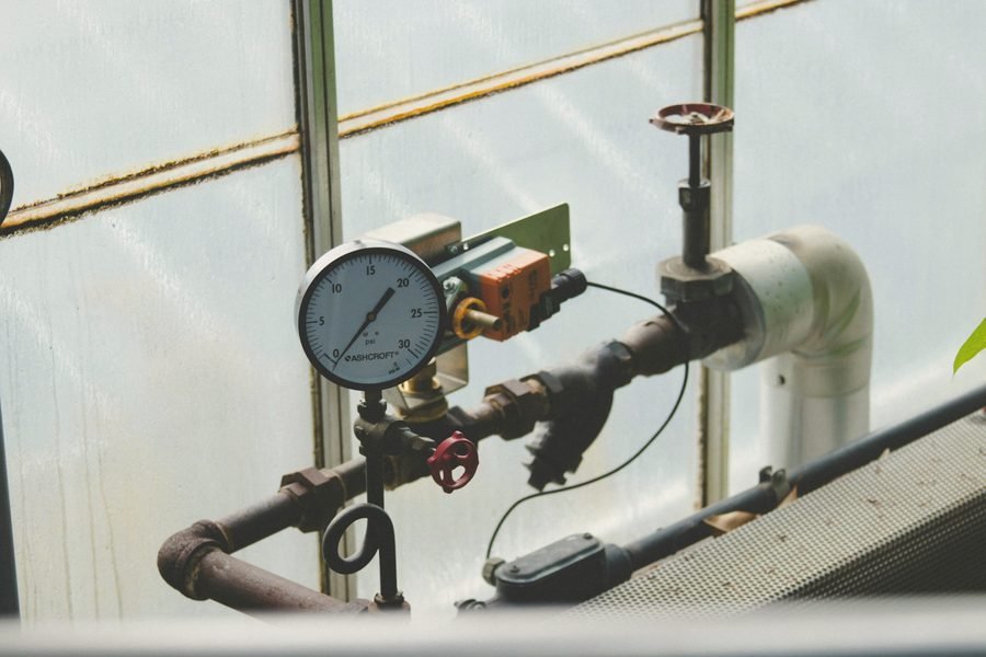 A pressure gauge and valves on a pipe run in front of a frosted window.