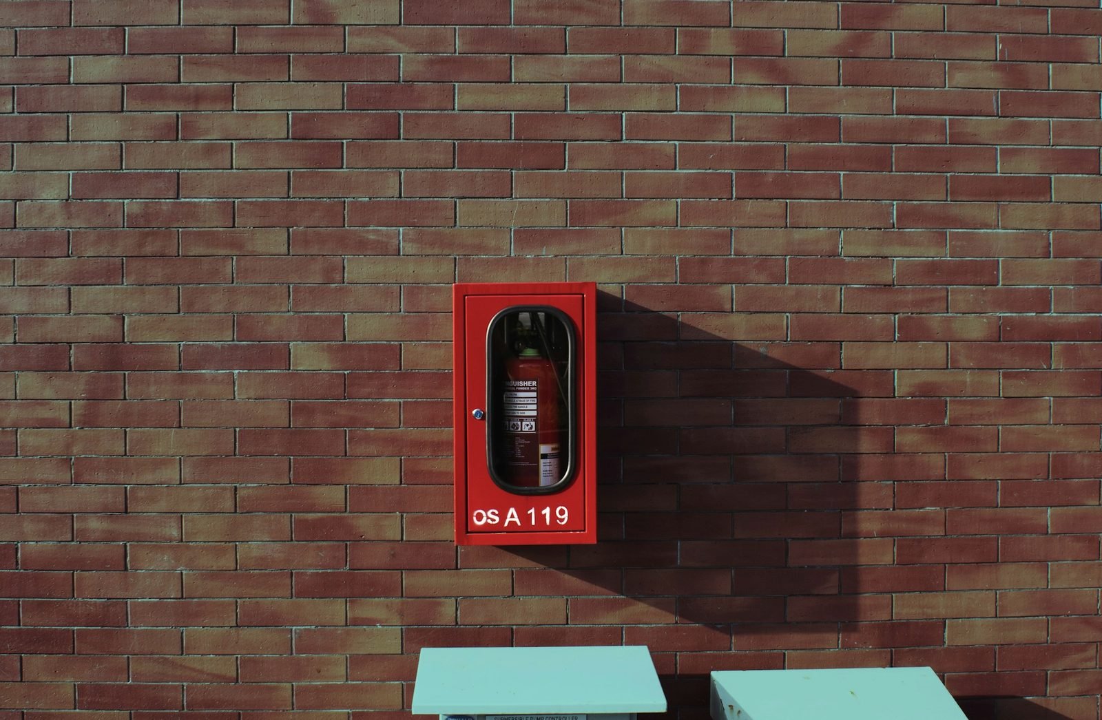 A red extinguisher cabinet with a glass window mounted on a brick wall in low afternoon sun.