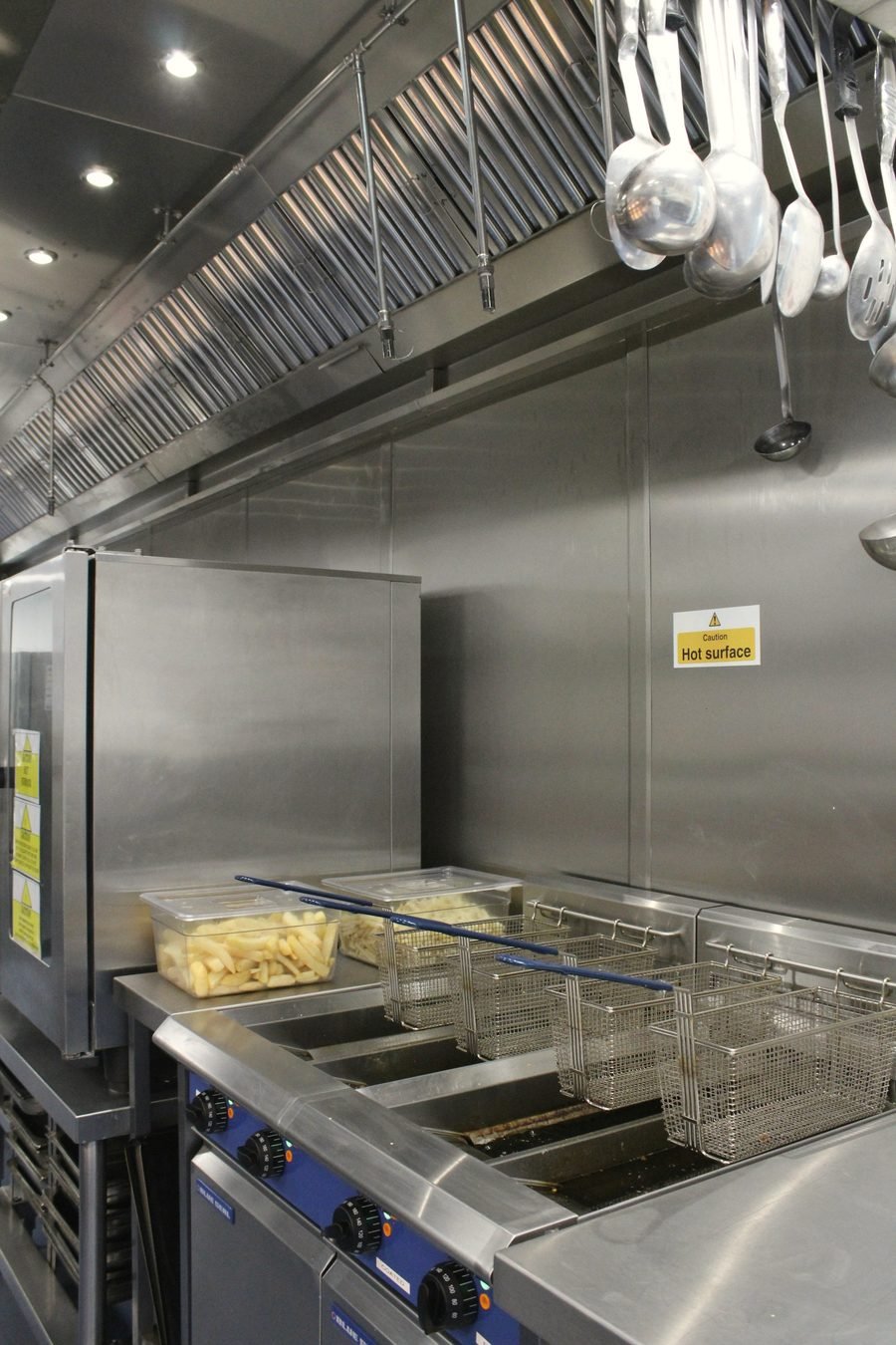 A bank of stainless deep fryers with wire baskets under a hood.