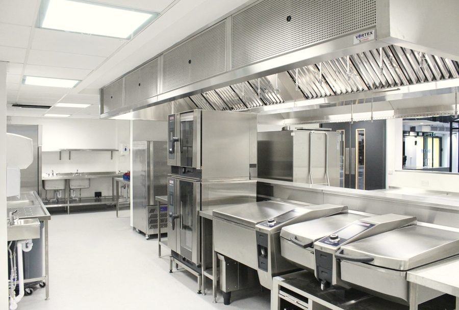 An empty stainless steel commercial kitchen with a long exhaust hood over the cooking line.