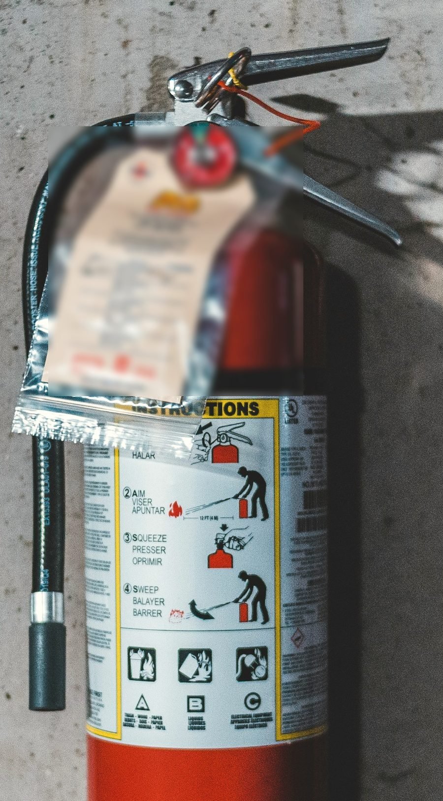 A wall-mounted extinguisher with a paper service tag hanging from the handle beside the gauge and pull pin.